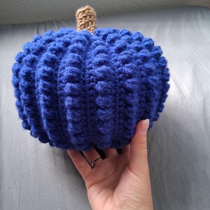 May include: A handmade, blue crochet pumpkin with a textured surface. The pumpkin has a brown stem and is held in a person's hand. This decorative item is perfect for fall and autumn decor.