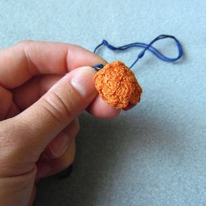 Puede incluir: Primer plano de un colgante de cuentas de ganchillo naranja con un cordón azul. La cuenta tiene una forma esférica texturizada. El colgante se sostiene en una mano sobre un fondo azul claro. La cuenta mide aproximadamente 2,5 cm de diámetro.