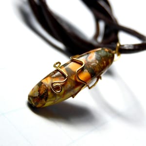 May include: A handcrafted necklace featuring a pendant with a speckled pattern in shades of brown, orange, and green. The pendant is wrapped with gold-colored wire and hangs from a dark brown leather cord. The necklace is set against a white background.