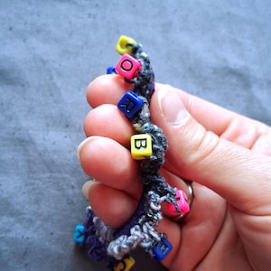 K&ouml;nnte beinhalten: Ein handgefertigtes Armband mit einem dunkelgrauen und violetten Webmuster. Das Armband hat bunte quadratische Perlen mit Buchstaben. Die Perlen sind rosa, gelb, blau und gr&uuml;n. Das Armband wird in einer Hand gehalten.