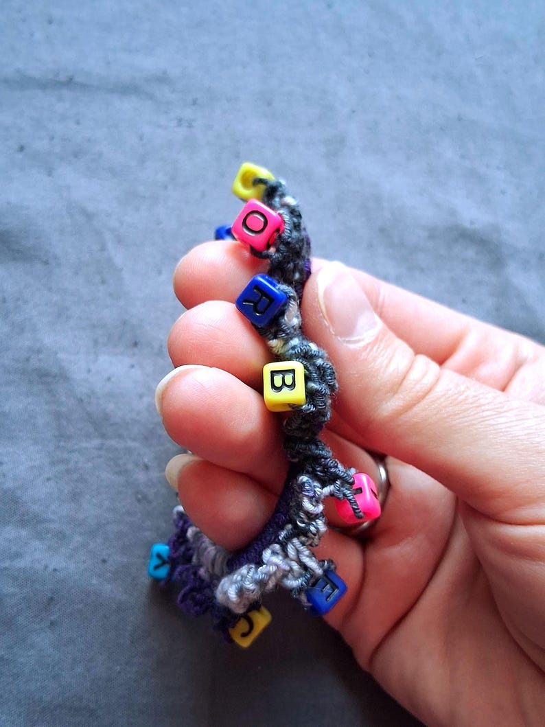 K&ouml;nnte beinhalten: Ein handgefertigtes Armband mit einem dunkelgrauen und violetten Webmuster. Das Armband hat bunte quadratische Perlen mit Buchstaben. Die Perlen sind rosa, gelb, blau und gr&uuml;n. Das Armband wird in einer Hand gehalten.