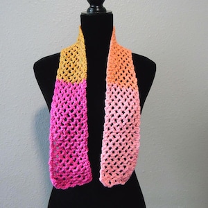 May include: A handmade crochet scarf in shades of yellow, pink, and orange. The scarf is displayed on a black mannequin. The open weave design is visible, and the colors transition from yellow to pink to orange.
