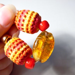 May include: A close-up of a colorful bracelet with red, orange, and yellow crocheted beads, amber-colored glass beads, and red spacer beads.