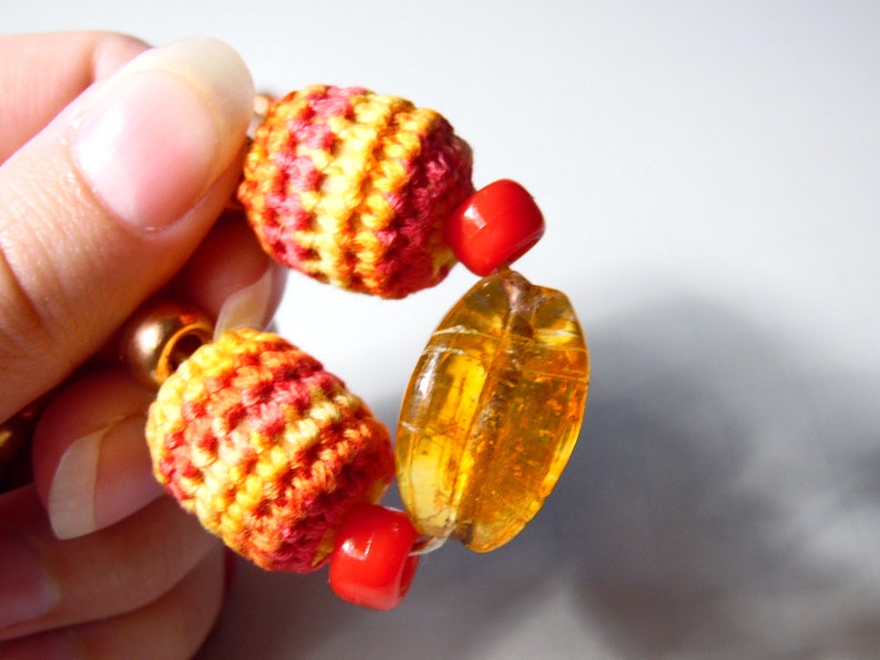 May include: A close-up of a colorful bracelet with red, orange, and yellow crocheted beads, amber-colored glass beads, and red spacer beads.