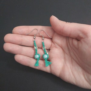 May include: A pair of teal green tassel earrings with silver hooks and a white bead in the center of each tassel.