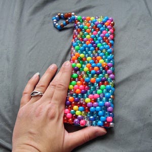 May include: A rectangular beaded phone case in a vibrant array of colors including blue, orange, pink, green, and purple. The case has a loop at the top and is held by a hand.