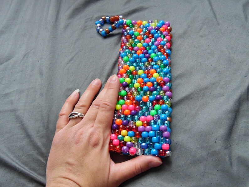 May include: A rectangular beaded phone case in a vibrant array of colors including blue, orange, pink, green, and purple. The case has a loop at the top and is held by a hand.
