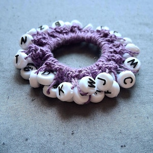 May include: A handmade purple scrunchie with white letter beads spelling out various letters. The scrunchie is made of a woven material and the beads are strung around the outside. The scrunchie is on a gray surface.