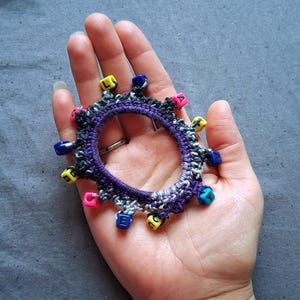 K&ouml;nnte beinhalten: Ein handgefertigtes H&auml;kelarmband in Lila, Grau und Wei&szlig;, verziert mit bunten Buchstabenperlen. Das Armband wird in einer Hand vor grauem Hintergrund gehalten. Die Perlen sind gelb, rosa, blau und gr&uuml;n.