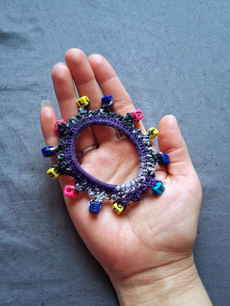 K&ouml;nnte beinhalten: Ein handgefertigtes H&auml;kelarmband in Lila, Grau und Wei&szlig;, verziert mit bunten Buchstabenperlen. Das Armband wird in einer Hand vor grauem Hintergrund gehalten. Die Perlen sind gelb, rosa, blau und gr&uuml;n.