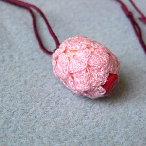 Puede incluir: Collar con colgante rosa hecho a mano. El colgante tiene forma ovalada con una cuenta roja en un extremo. El cordón del collar es de color burdeos oscuro. El colgante está hecho de hilo suave y texturizado.