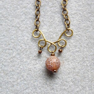 May include: A handcrafted necklace featuring a gold-toned chain and decorative elements. The focal point is a round, orange and white patterned bead pendant. Small, dark brown beads dangle from the design.