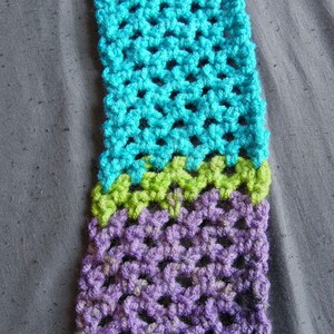 May include: A close-up of a crocheted scarf in shades of turquoise, green, and purple. The handmade scarf features a textured, open weave design. The color transitions are clearly defined, creating a visually appealing pattern. The scarf is displayed on a gray surface.