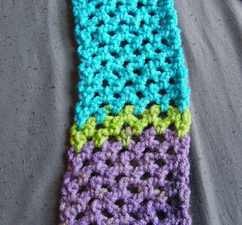 May include: A close-up of a crocheted scarf in shades of turquoise, green, and purple. The handmade scarf features a textured, open weave design. The color transitions are clearly defined, creating a visually appealing pattern. The scarf is displayed on a gray surface.