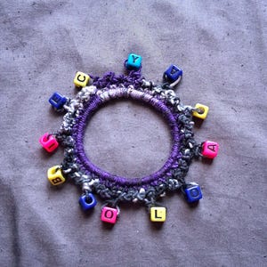 K&ouml;nnte beinhalten: Ein handgefertigtes Armband mit einem lila, schwarzen und wei&szlig;en geh&auml;kelten Band. Das Armband ist mit bunten quadratischen Perlen verziert, die jeweils einen einzelnen Buchstaben zeigen. Die Perlen sind in Rosa-, Gelb-, Blau- und Gr&uuml;nt&ouml;nen gehalten.