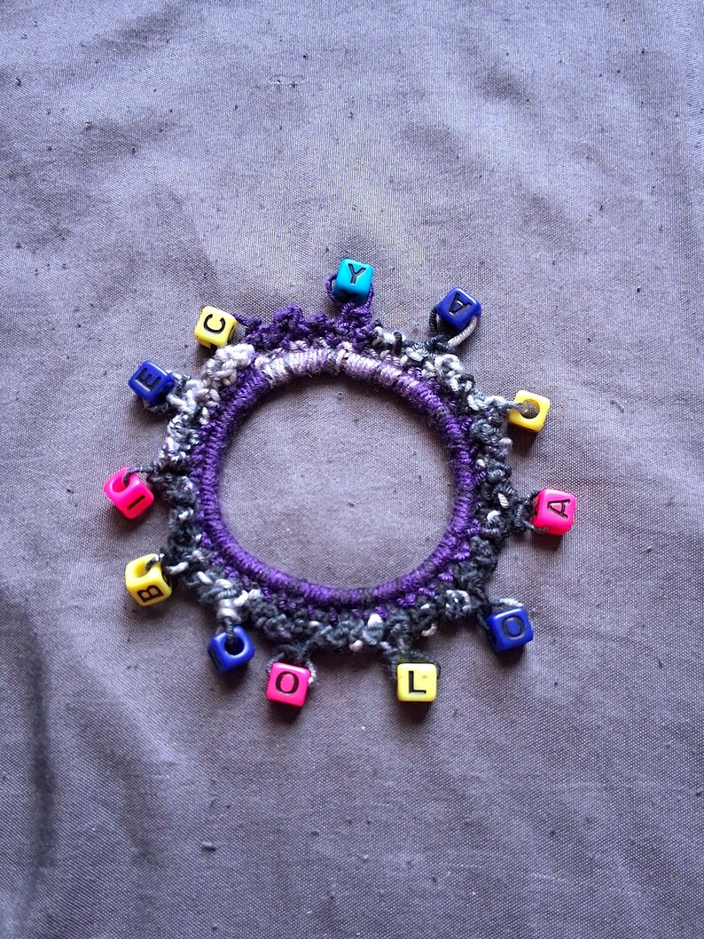 K&ouml;nnte beinhalten: Ein handgefertigtes Armband mit einem lila, schwarzen und wei&szlig;en geh&auml;kelten Band. Das Armband ist mit bunten quadratischen Perlen verziert, die jeweils einen einzelnen Buchstaben zeigen. Die Perlen sind in Rosa-, Gelb-, Blau- und Gr&uuml;nt&ouml;nen gehalten.