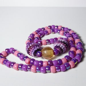 Puede incluir: Un collar de cuentas de color morado y rosa con dos cuentas de ganchillo y una cuenta de vidrio transparente.