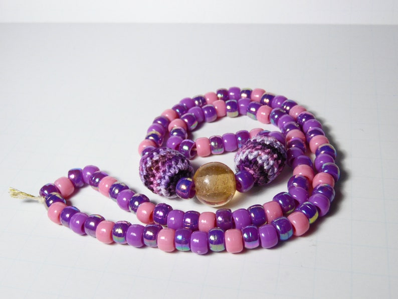 Puede incluir: Un collar de cuentas de color morado y rosa con dos cuentas de ganchillo y una cuenta de vidrio transparente.