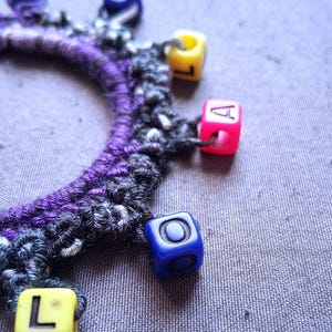 K&ouml;nnte beinhalten: Ein handgefertigtes Armband mit einem lila-grauen H&auml;kelband. Das Armband ist mit bunten quadratischen Perlen verziert, darunter gelbe, rosa und blaue, die jeweils einen Buchstaben oder ein Symbol zeigen. Der Hintergrund ist ein strukturierter grauer Stoff.