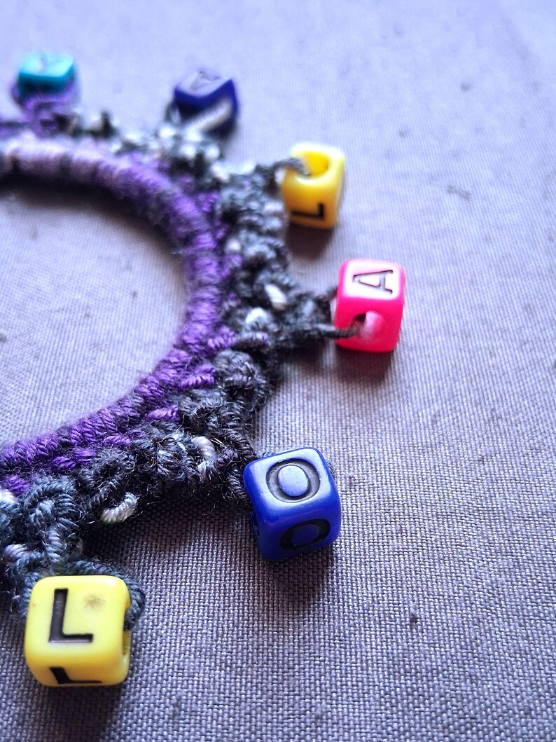K&ouml;nnte beinhalten: Ein handgefertigtes Armband mit einem lila-grauen H&auml;kelband. Das Armband ist mit bunten quadratischen Perlen verziert, darunter gelbe, rosa und blaue, die jeweils einen Buchstaben oder ein Symbol zeigen. Der Hintergrund ist ein strukturierter grauer Stoff.