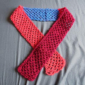 May include: A handmade crochet scarf in shades of red, pink, and blue. The scarf is made with a lacy stitch, and the colors transition from red to pink to blue. The scarf is laid out on a gray surface.
