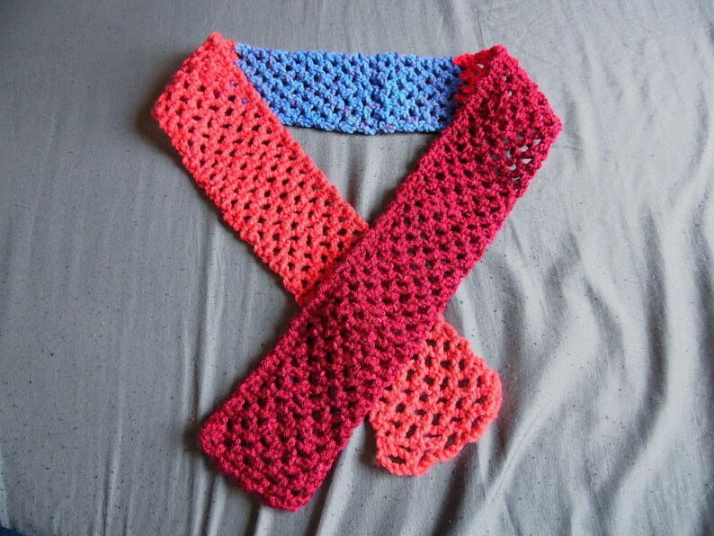 May include: A handmade crochet scarf in shades of red, pink, and blue. The scarf is made with a lacy stitch, and the colors transition from red to pink to blue. The scarf is laid out on a gray surface.
