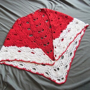 May include: A handmade, red and white crochet shawl. The shawl features a detailed openwork design with a scalloped white border. The red portion has a repeating pattern of circular cutouts. The shawl is displayed on a gray surface.