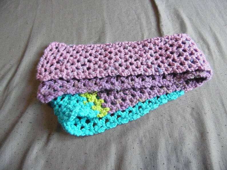 May include: A handmade crochet headband in shades of pink, purple, and turquoise. The headband is folded, revealing layers of textured yarn. A small section of lime green yarn is visible within the turquoise layer.