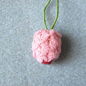 Puede incluir: Colgante rosa de ganchillo hecho a mano con una cuenta roja en la base, suspendido por un cordón verde. El colgante tiene un aspecto texturizado y en capas, similar a un capullo floral. El fondo es de tela gris claro.