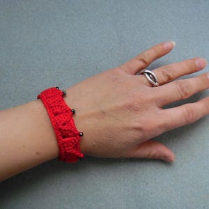 May include: A red crocheted bracelet with black beads. The bracelet is worn on a person's wrist.