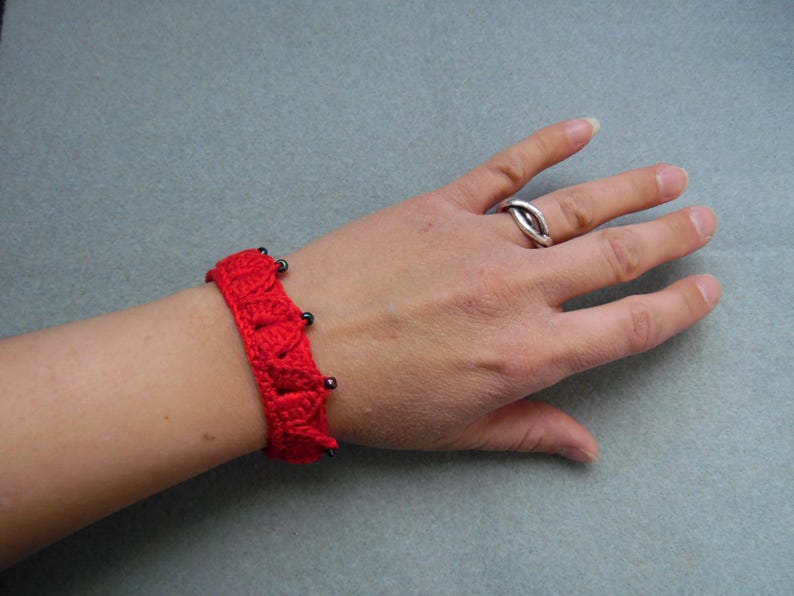 May include: A red crocheted bracelet with black beads. The bracelet is worn on a person's wrist.