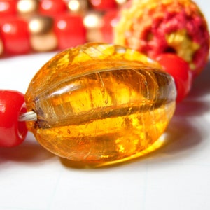 May include: A close-up of a necklace with red, gold, and orange beads. The necklace features a large, clear, amber-colored glass bead with a striated pattern.