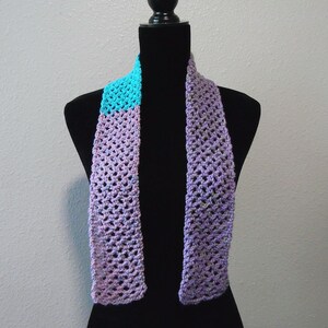 May include: A handmade crochet scarf in shades of purple and turquoise. The scarf is displayed on a black mannequin. The open weave design and color combination create a unique accessory. The scarf is a stylish addition to any wardrobe.