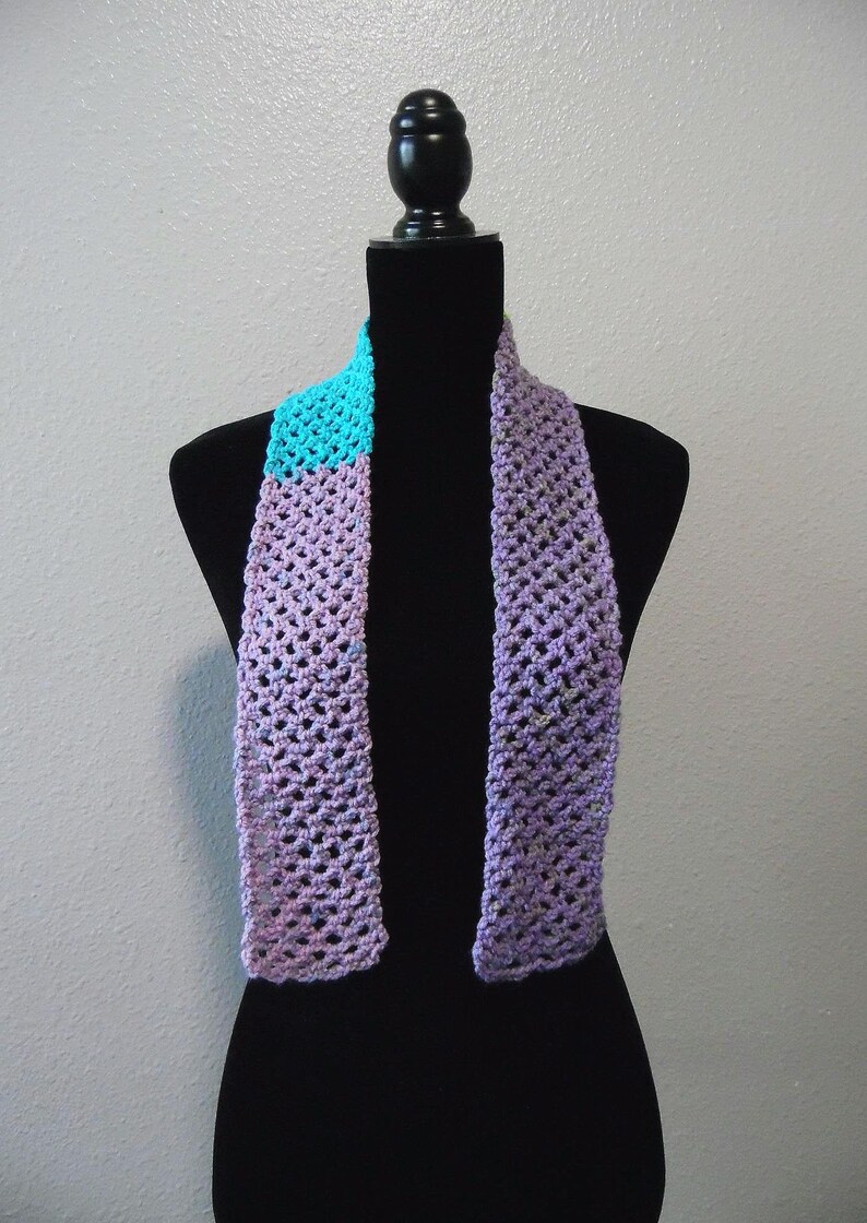 May include: A handmade crochet scarf in shades of purple and turquoise. The scarf is displayed on a black mannequin. The open weave design and color combination create a unique accessory. The scarf is a stylish addition to any wardrobe.