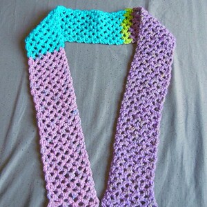 May include: A handmade crochet scarf in shades of blue, green, and purple. The scarf features an open, lacy stitch pattern. The top section is turquoise, transitioning to green, and then to a lavender-purple color. The ends have a scalloped edge.