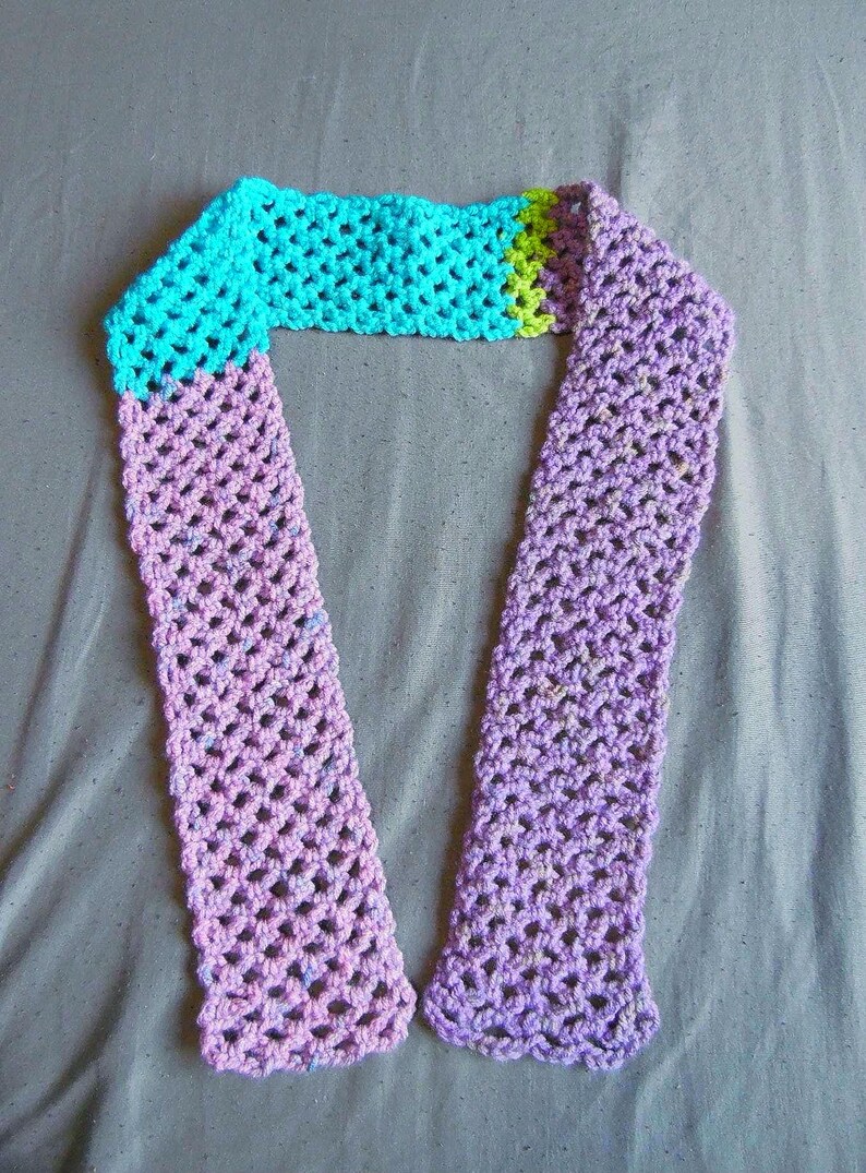 May include: A handmade crochet scarf in shades of blue, green, and purple. The scarf features an open, lacy stitch pattern. The top section is turquoise, transitioning to green, and then to a lavender-purple color. The ends have a scalloped edge.