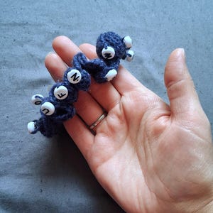 May include: Two navy blue crocheted creatures with white accents and lettered buttons. The handmade figures have a whimsical design, with the letters "F", "N", and "W" visible on the buttons. The creatures are displayed against a gray background.