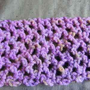 May include: Close-up of a crocheted item in shades of purple, with hints of tan and blue. The openwork design creates a textured pattern. The item is likely a scarf or a similar accessory.