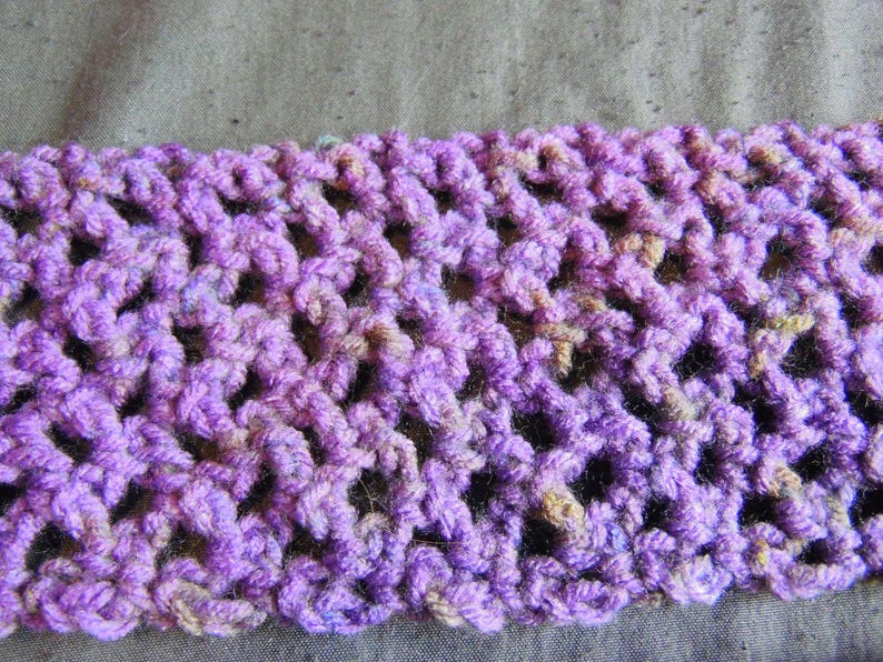 May include: Close-up of a crocheted item in shades of purple, with hints of tan and blue. The openwork design creates a textured pattern. The item is likely a scarf or a similar accessory.