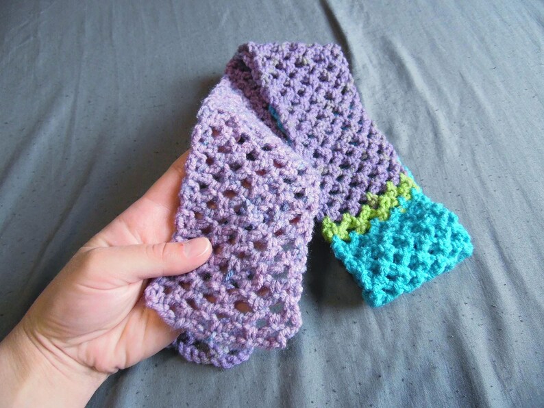 May include: A hand-crocheted scarf in shades of purple, with a turquoise and green border. The scarf is made with a lacy stitch, and the colors blend together. The scarf is approximately 60 inches long.