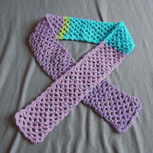 May include: A handmade crochet scarf in shades of purple, teal, and green. The scarf is made with a open, lacy stitch. The scarf is a long, rectangular shape, with a color block design. The scarf is a cozy accessory.