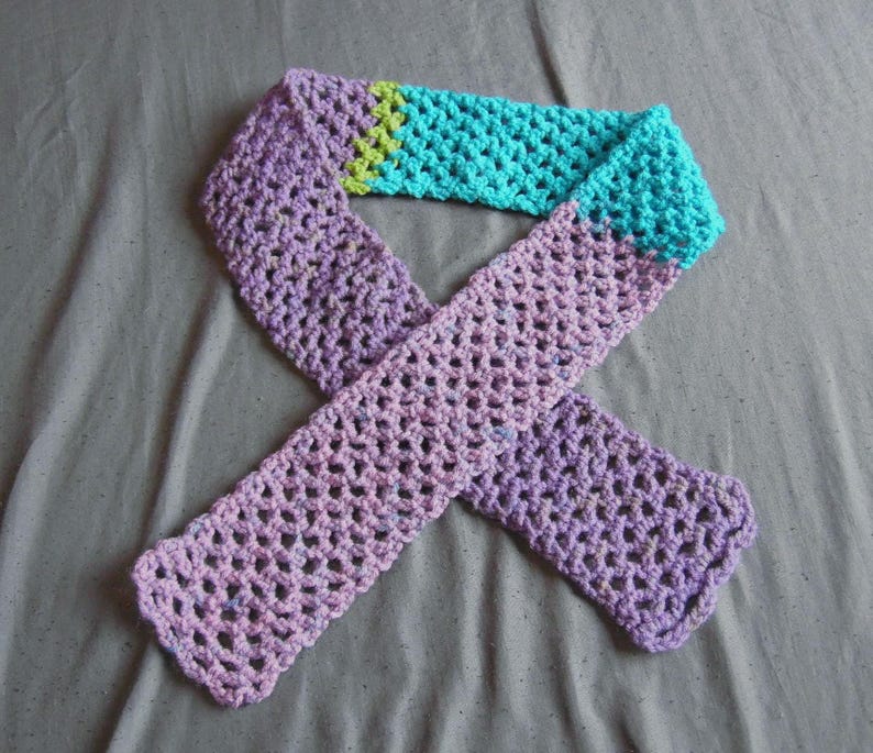 May include: A handmade crochet scarf in shades of purple, teal, and green. The scarf is made with a open, lacy stitch. The scarf is a long, rectangular shape, with a color block design. The scarf is a cozy accessory.