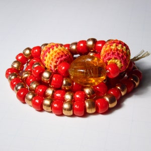 May include: A red and gold beaded necklace with crocheted orange, yellow, and red beads. The necklace is coiled up on a white surface.