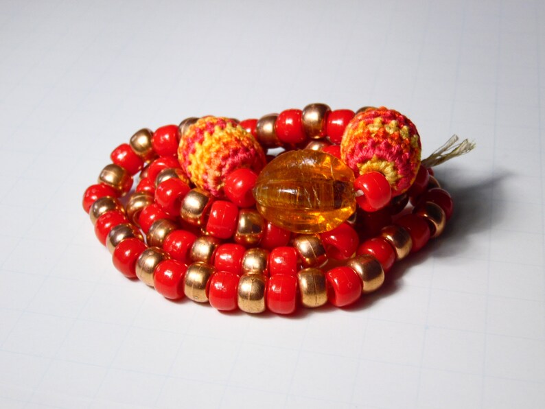 May include: A red and gold beaded necklace with crocheted orange, yellow, and red beads. The necklace is coiled up on a white surface.
