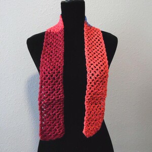 May include: A handmade, open-weave scarf in shades of red and orange, displayed on a black mannequin. The scarf's gradient color transitions from a deep red to a vibrant orange. The open weave design adds texture and visual interest to the accessory.