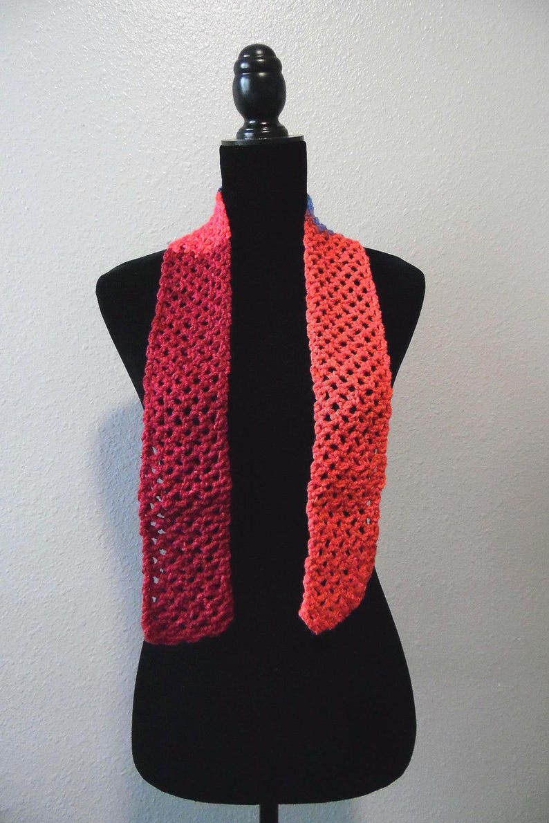 May include: A handmade, open-weave scarf in shades of red and orange, displayed on a black mannequin. The scarf's gradient color transitions from a deep red to a vibrant orange. The open weave design adds texture and visual interest to the accessory.