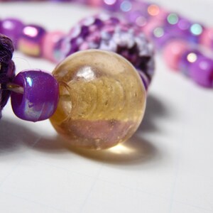 Puede incluir: Primer plano de una cuenta de ganchillo de color morado y blanco, una cuenta iridiscente morada y una cuenta de vidrio transparente con un patr&oacute;n de espiral.