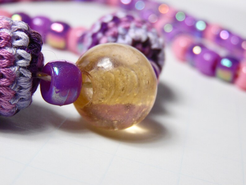 Puede incluir: Primer plano de una cuenta de ganchillo de color morado y blanco, una cuenta iridiscente morada y una cuenta de vidrio transparente con un patr&oacute;n de espiral.