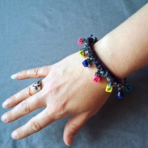 K&ouml;nnte beinhalten: Ein handgefertigtes H&auml;kelarmband in Schwarz und Wei&szlig; mit bunten quadratischen Perlen. Das Armband ist mit kleinen, quadratischen Perlen in verschiedenen Farben verziert, die jeweils einen Buchstaben zeigen. Ein Silberring befindet sich an der Hand.