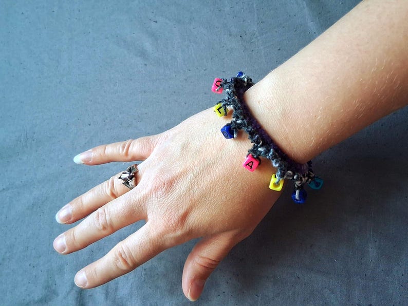 K&ouml;nnte beinhalten: Ein handgefertigtes H&auml;kelarmband in Schwarz und Wei&szlig; mit bunten quadratischen Perlen. Das Armband ist mit kleinen, quadratischen Perlen in verschiedenen Farben verziert, die jeweils einen Buchstaben zeigen. Ein Silberring befindet sich an der Hand.